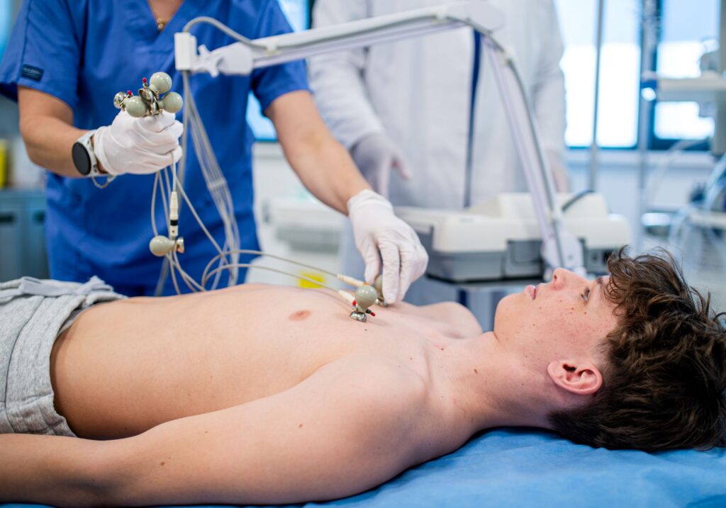 Leżący nastolatek oraz lekarz z pielęgniarką wykonujący badanie EKG – ilustracja dotycząca elektrokardiografii dzieci w Centrum Medycznym neoMedica w Poznaniu.